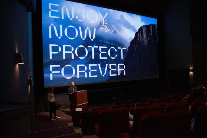Speaker presents on stage with Enjoy Now Protect Forever message on large screen in cin...