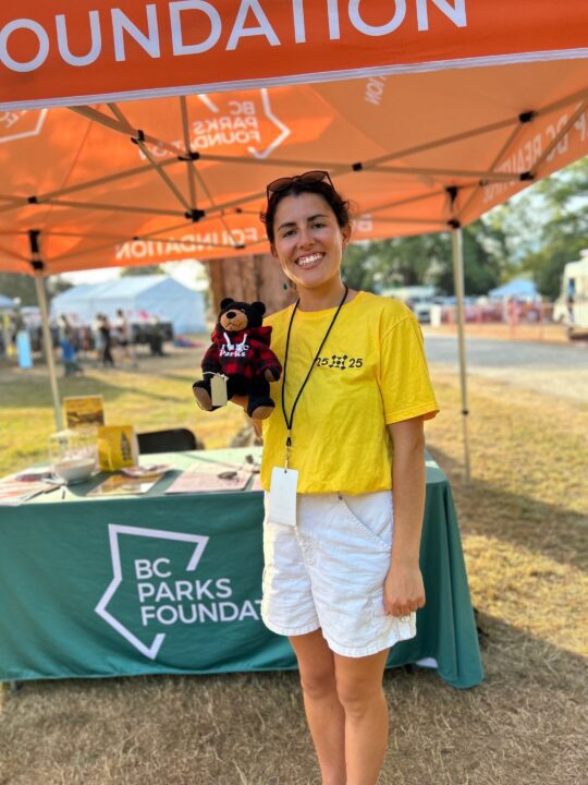 Person holding a bear plush at BC Parks Foundation booth, outdoor event; promoting park...