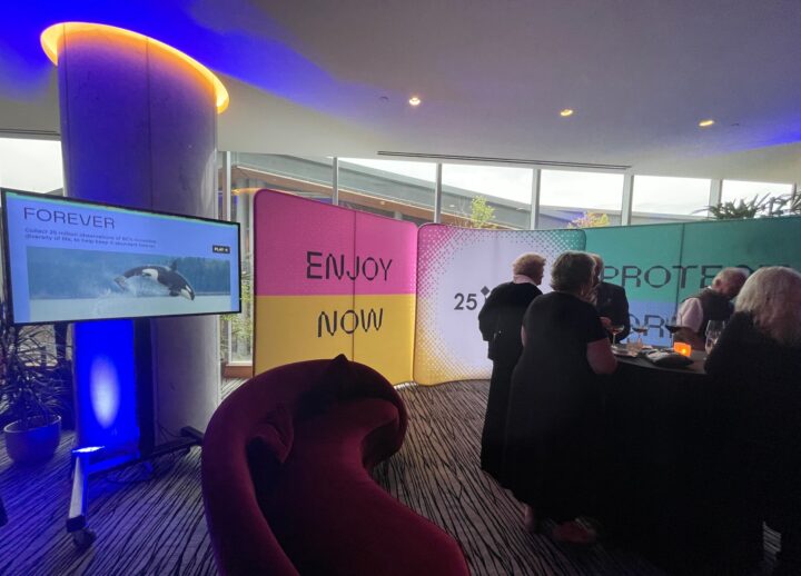 Event gathering with colorful Enjoy Now, Protect Forever banners, showcasing a TV scree...