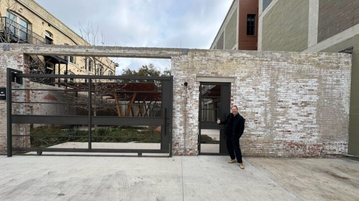 Corey Gutch smiling and giving thumbs up in front of the industrial gate and brick building headquarters of Lake Flato in San Antonio, Texas.