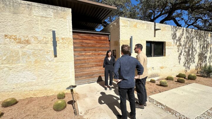 Michael Ellsworth, Evan Morris, and Vicki Yuan stand outside Lake Flato's Olmos Park Residence a modern stone house with wooden door, discussing architecture in a sunny setting.