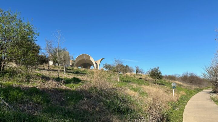 Unique architectural structure on a grassy hill under a clear blue sky, bordered by a w...