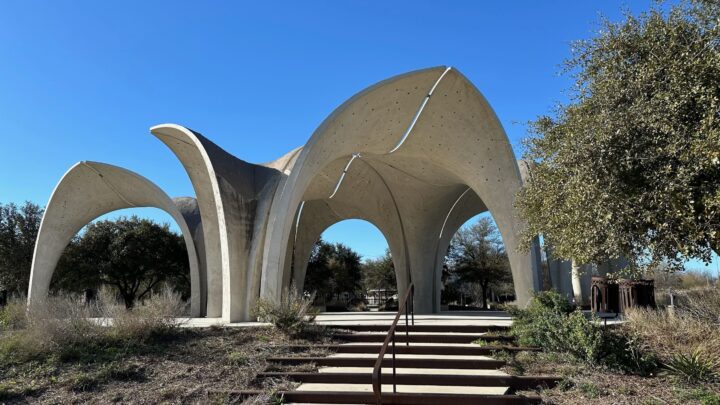 Futuristic concrete arch structure under clear blue sky, surrounded by trees and greenery.