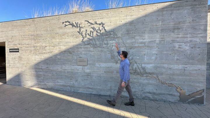 Man points at artistic map on concrete wall, sunlight casts shadow, restroom sign visib...