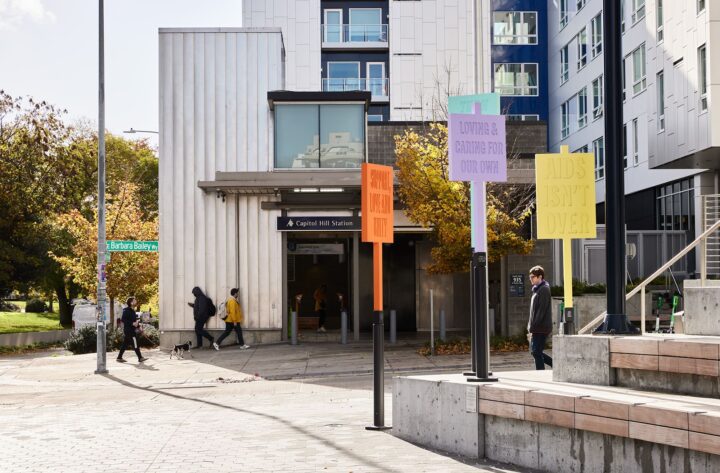 People passing Capitol Hill Station entrance in autumn. Colorful signs display messages...