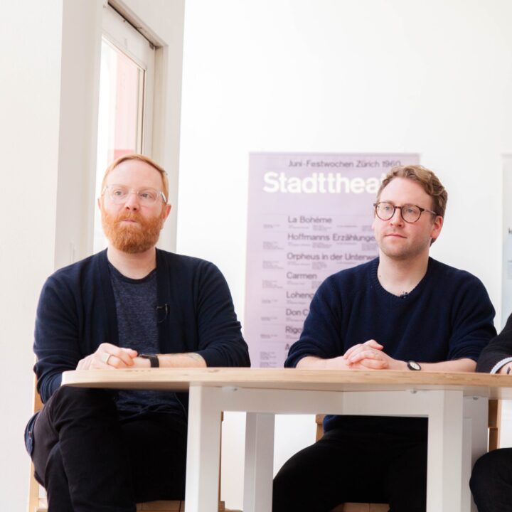 Three men sitting at a table in a modern room with theater posters behind them.