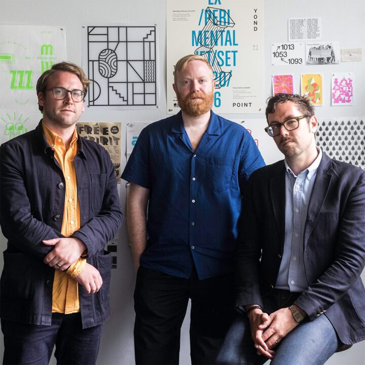 Three men pose in front of creative posters on a wall, showcasing a modern design studio atmosphere.