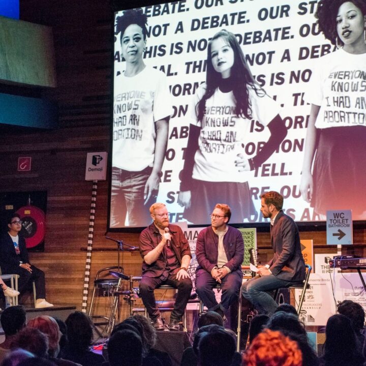 Panel discussion on stage with large abortion rights image display in background, featuring three seated speakers and attentive audience.