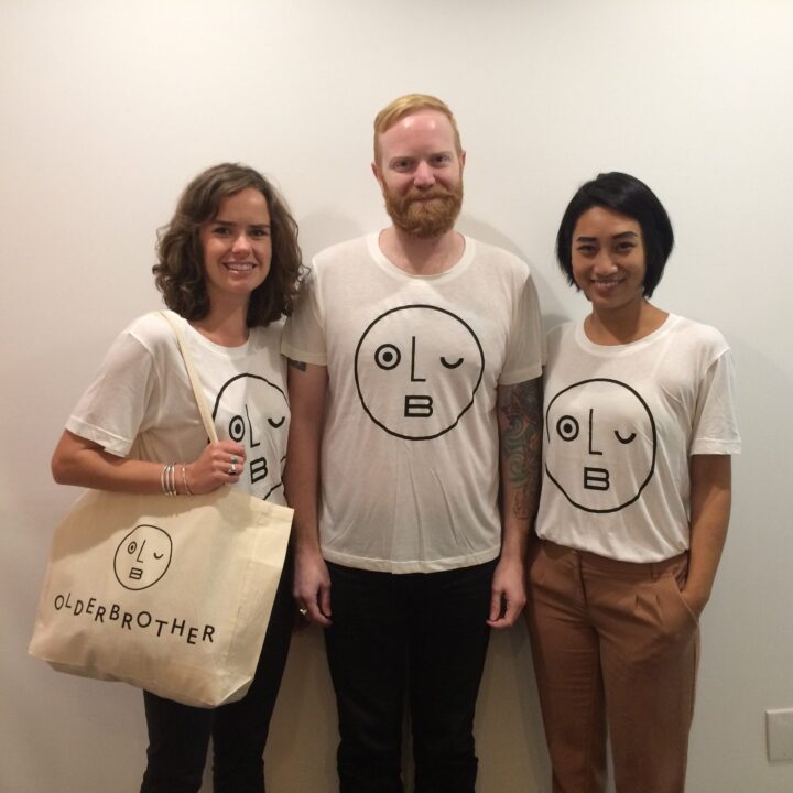 Three people wearing matching Olderbrother t-shirts and a tote bag, standing against a white wall, smiling and posing.