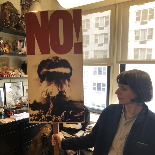 Person holding a protest sign with a mushroom cloud and large NO! text, standing in an office environment.