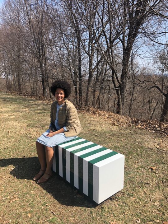 Smiling person sitting on a striped bench in a park during early spring, surrounded by bare trees and sunny skies.
