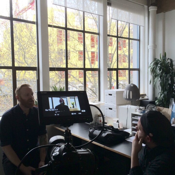 Man being filmed in an office setting with large windows, camera and monitor visible; conversation setting.