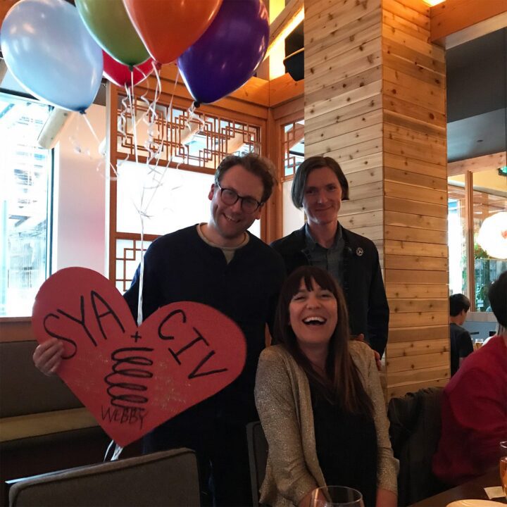 Three people laughing at a party with colorful balloons and a red heart-shaped sign inside a wooden-paneled room.