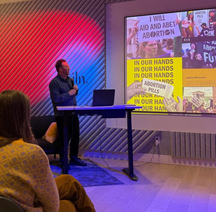 Speaker presents on abortion rights and access to abortion pills at an indoor event. Attendees listen to the presentation.