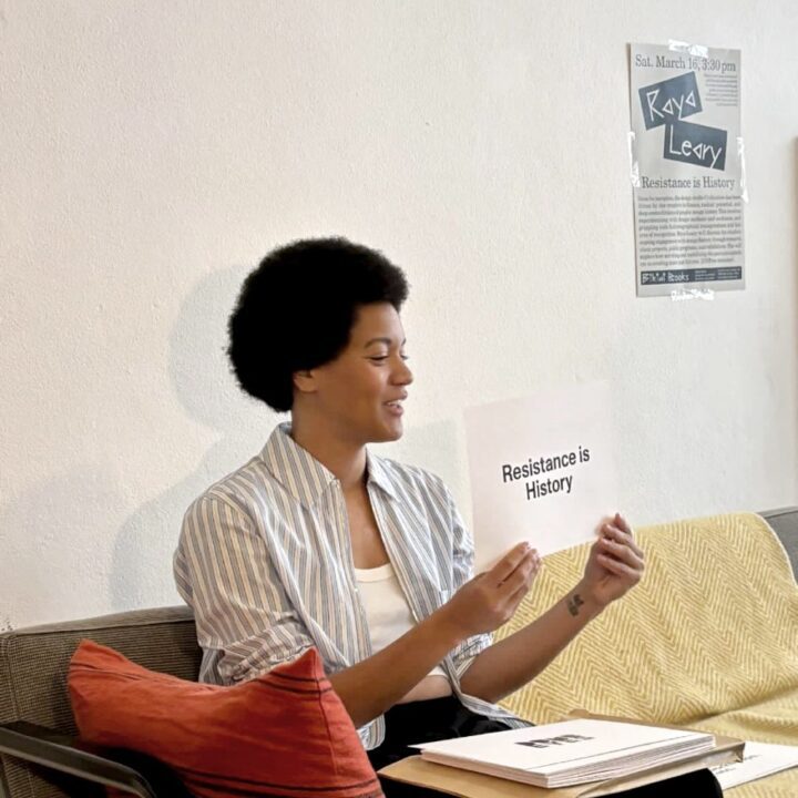 Raya Leary holding a sign saying Resistance is History, sitting on a couch with a poster in the background, during her lecture at Bikini Books in Porto, Portugal.