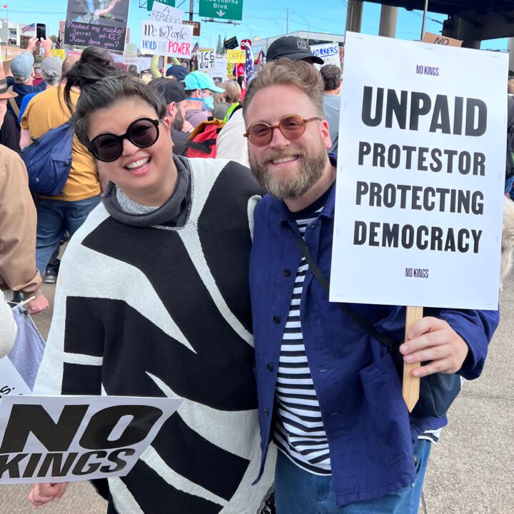 Two people smiling, holding a sign reading Unpaid Protestor Protecting Democracy at a peaceful rally.